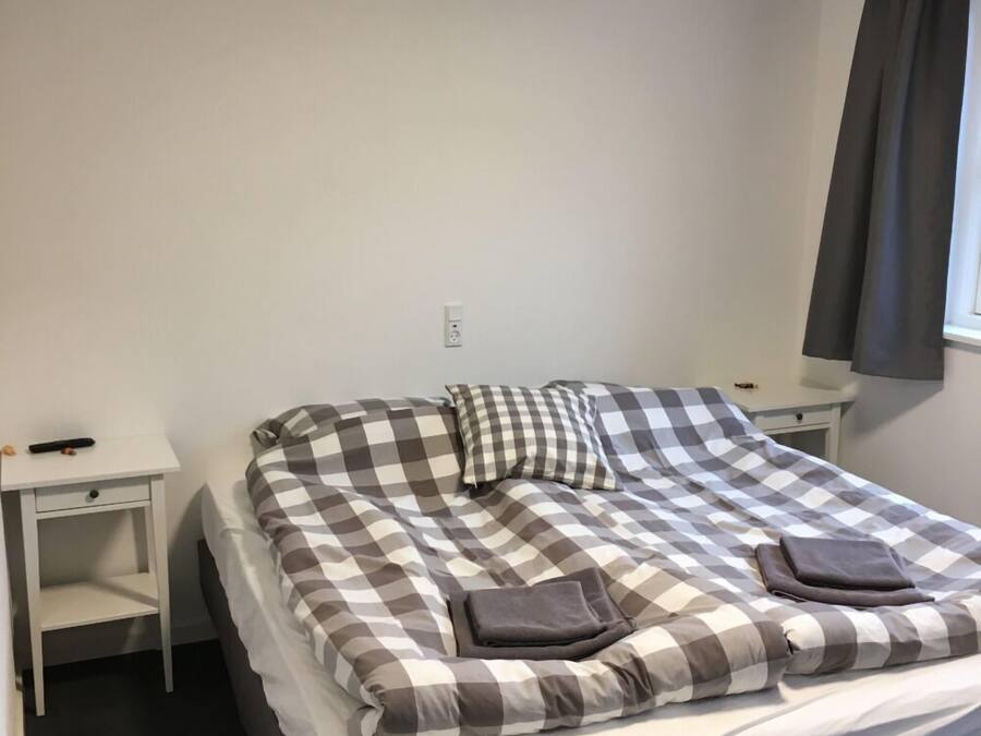 A single bed with a checkered bedspread, two pillows, and two folded towels. There is a white nightstand to the left and a white dresser to the right.