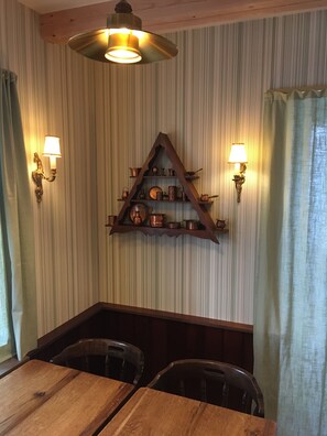 A dining area with a wooden table and chairs, a triangular shelf with copper cookware, and wall-mounted lamps. - Hotel Osted (Lejre)