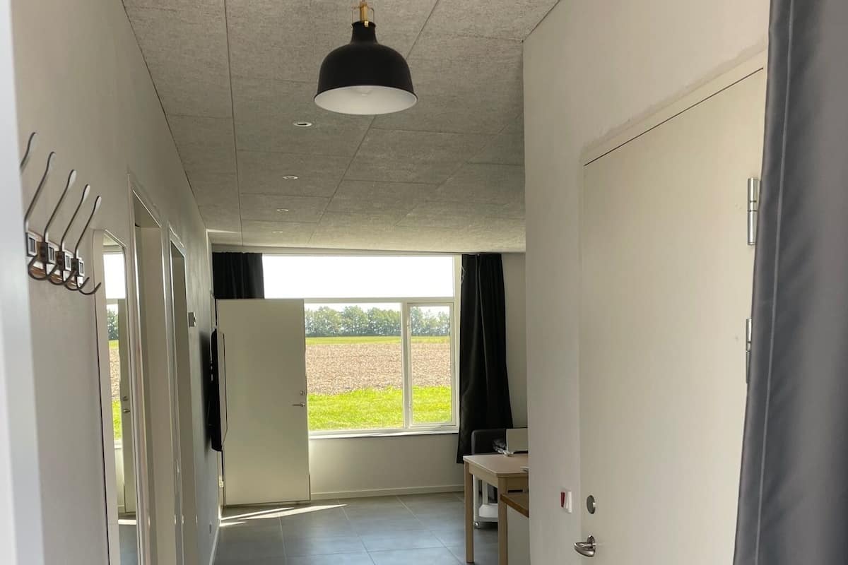 A hallway with a tiled floor, a hanging pendant light, a coat rack, and a door leading to a room with a view of a green field.