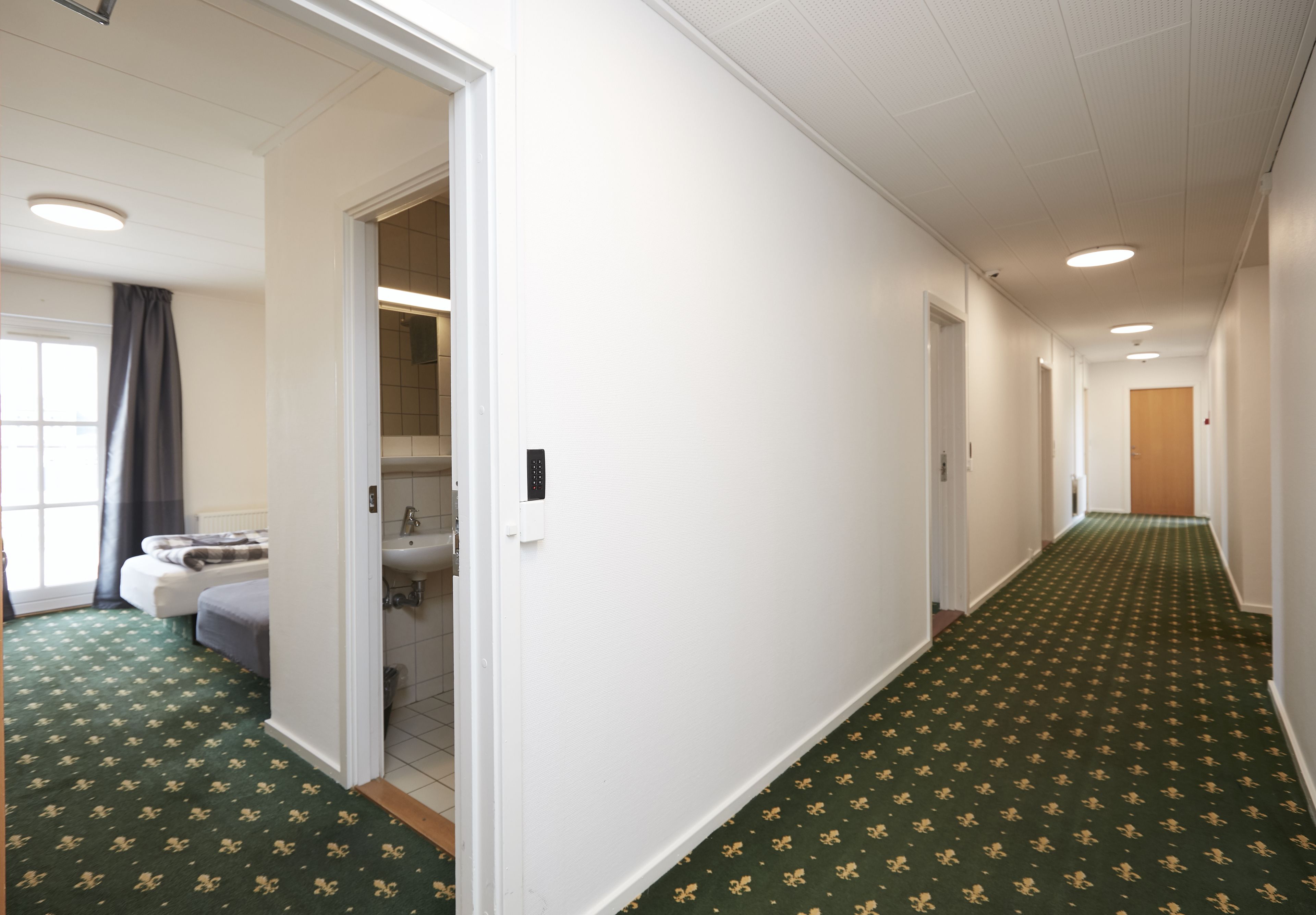A hotel corridor with a green patterned carpet, white walls, and a visible bathroom with a sink and toilet.