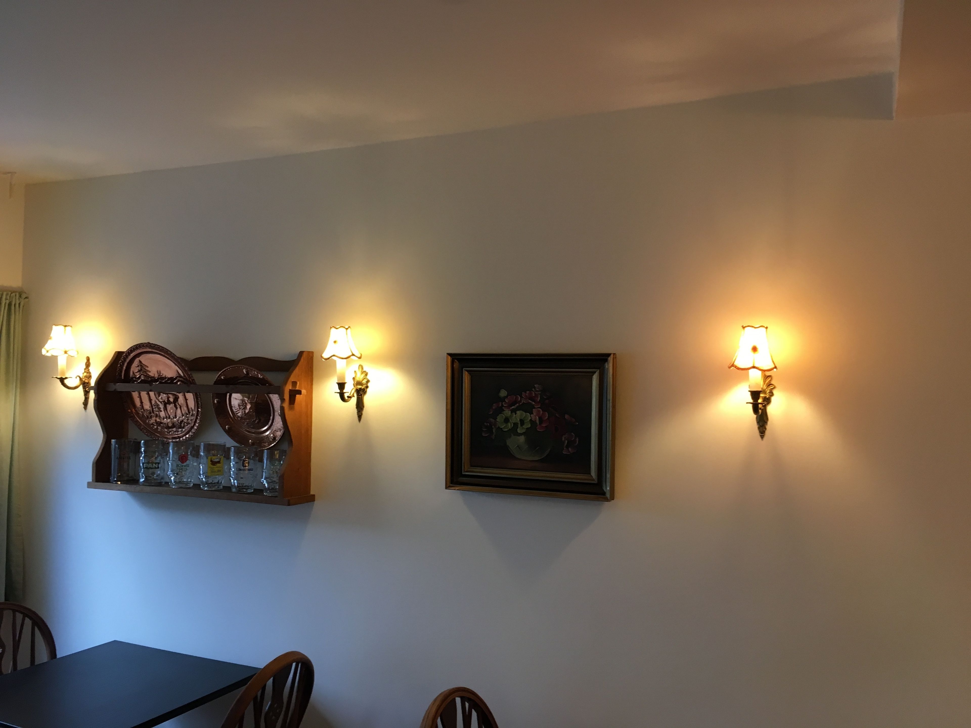 A dining area with a table, chairs, wall-mounted light fixtures, and decorative items including framed artwork and a shelf with glassware.
