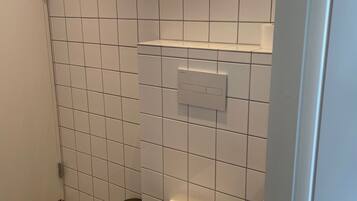 Um banheiro pequeno e limpo, com azulejos brancos, vaso sanitário, janela e área de chuveiro.