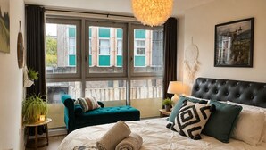 A bedroom with a large bed, a tufted headboard, a hanging light fixture, a view of buildings through large windows, and a small side table with a plant. - Lovely Bohemian Apartment in Heart of City Life (Glasgow)
