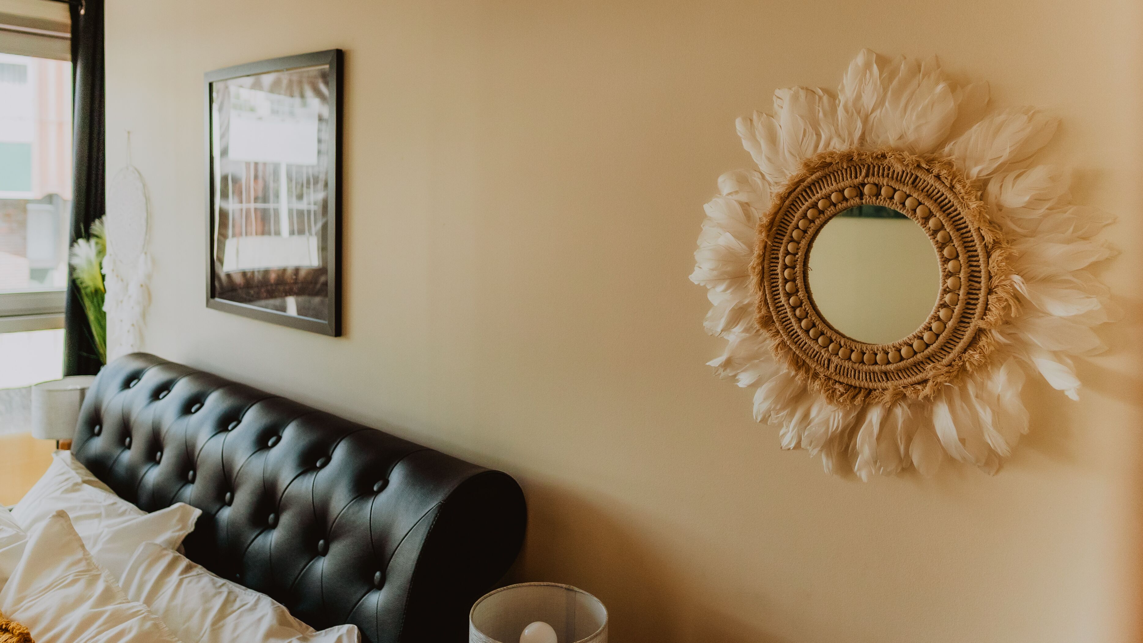A bedroom with a tufted headboard, a wall-mounted mirror with a feathered frame, and a framed picture above the headboard.