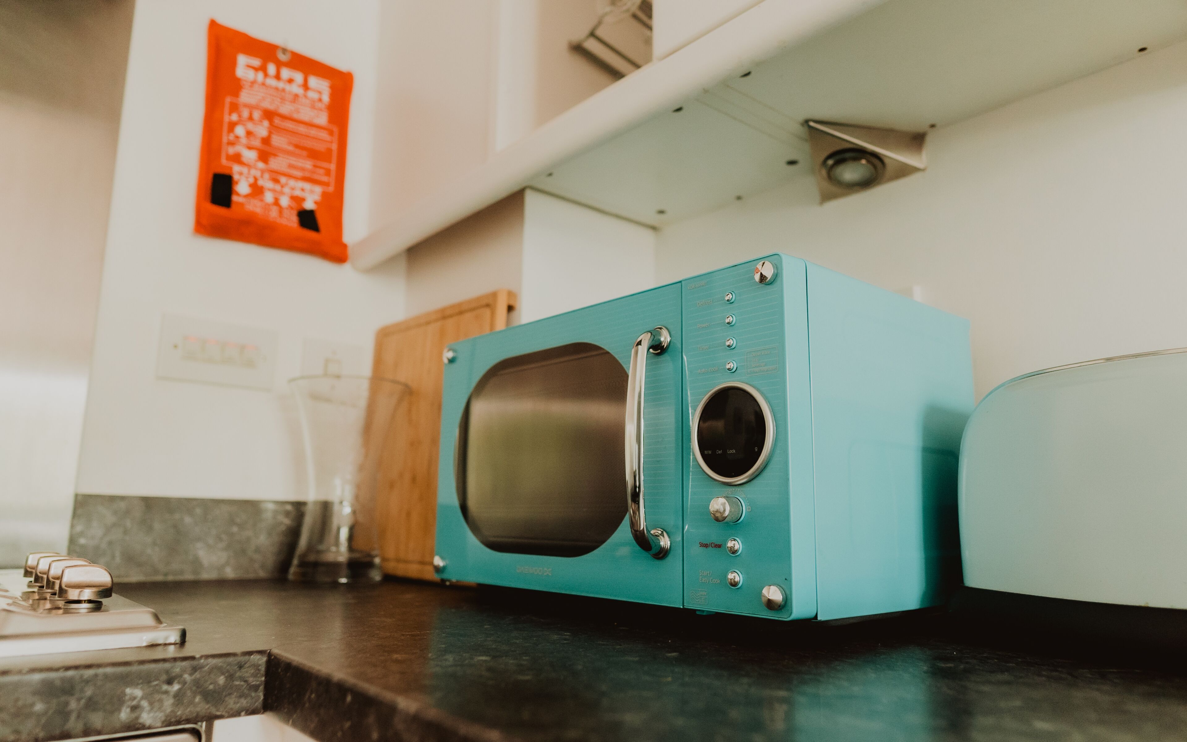 Un microondas color turquesa sobre una encimera de cocina con un mueble de madera y un vaso al fondo.