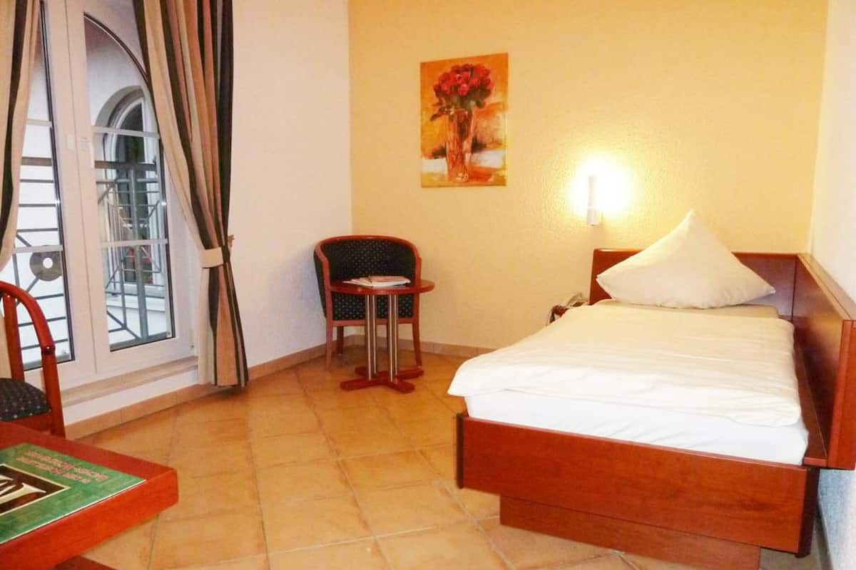 Habitación de hotel con cama, una silla, una mesita, un cuadro y una ventana con cortinas.