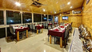 A restaurant with wooden walls and ceiling, multiple tables with red tablecloths, and a television mounted on the wall. - Hotel Monarch Residency (Manali)