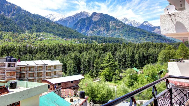 A mountainous landscape with a view of snow-capped peaks and dense forest. There are buildings with multiple floors and a satellite dish visible. - Hotel Monarch Residency (Manali)