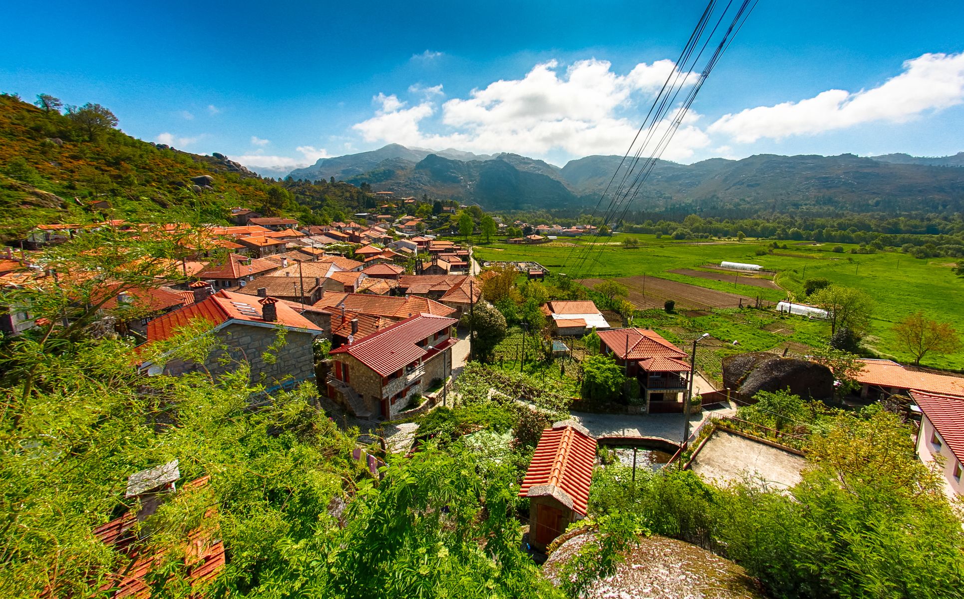A village with traditional red-tiled roofs, surrounded by lush greenery and mountains in the background.