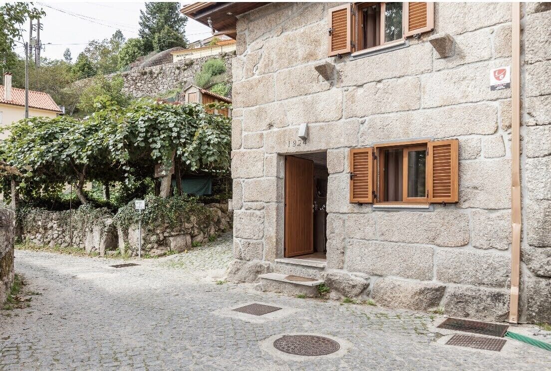 A stone building with a wooden door and shutters, a cobblestone street, and a stone wall with vegetation.