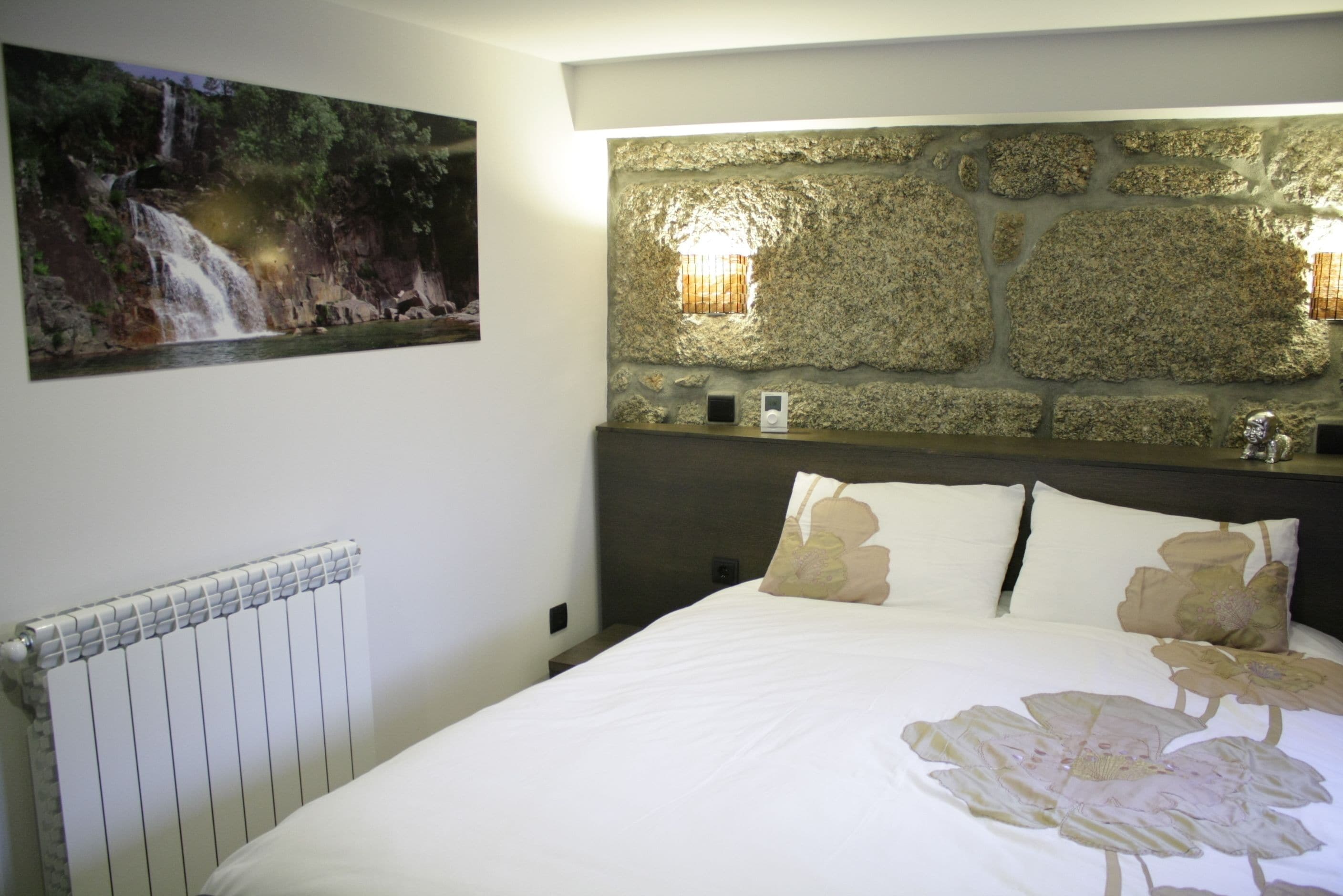 Un dormitorio con una cama grande, una pared de piedra, un cuadro con una cascada y un radiador.