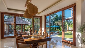 Un comedor amplio con una gran mesa de madera, sillas cómodas y una moderna lámpara de araña. Amplios ventanales ofrecen vistas a un jardín exuberante y una zona de piscina.