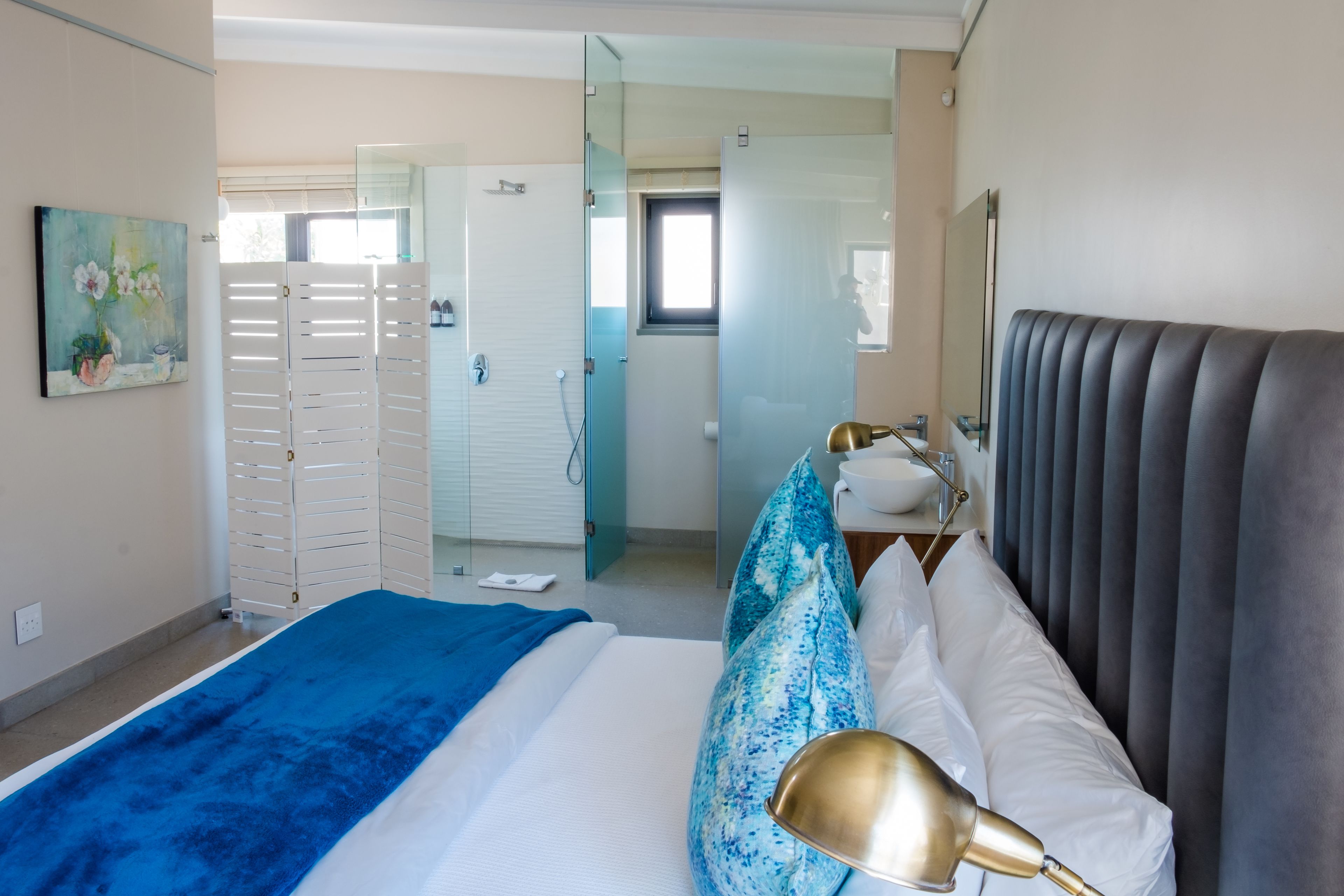 A modern hotel room with a bed, a bedside lamp, a bathroom with a glass door, and a painting on the wall.