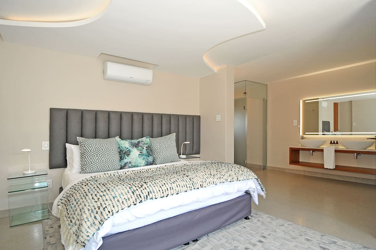 Quarto moderno com cama grande, mesa de cabeceira, criado-mudo, banco embutido, espelho grande e banheiro com pia e chuveiro.