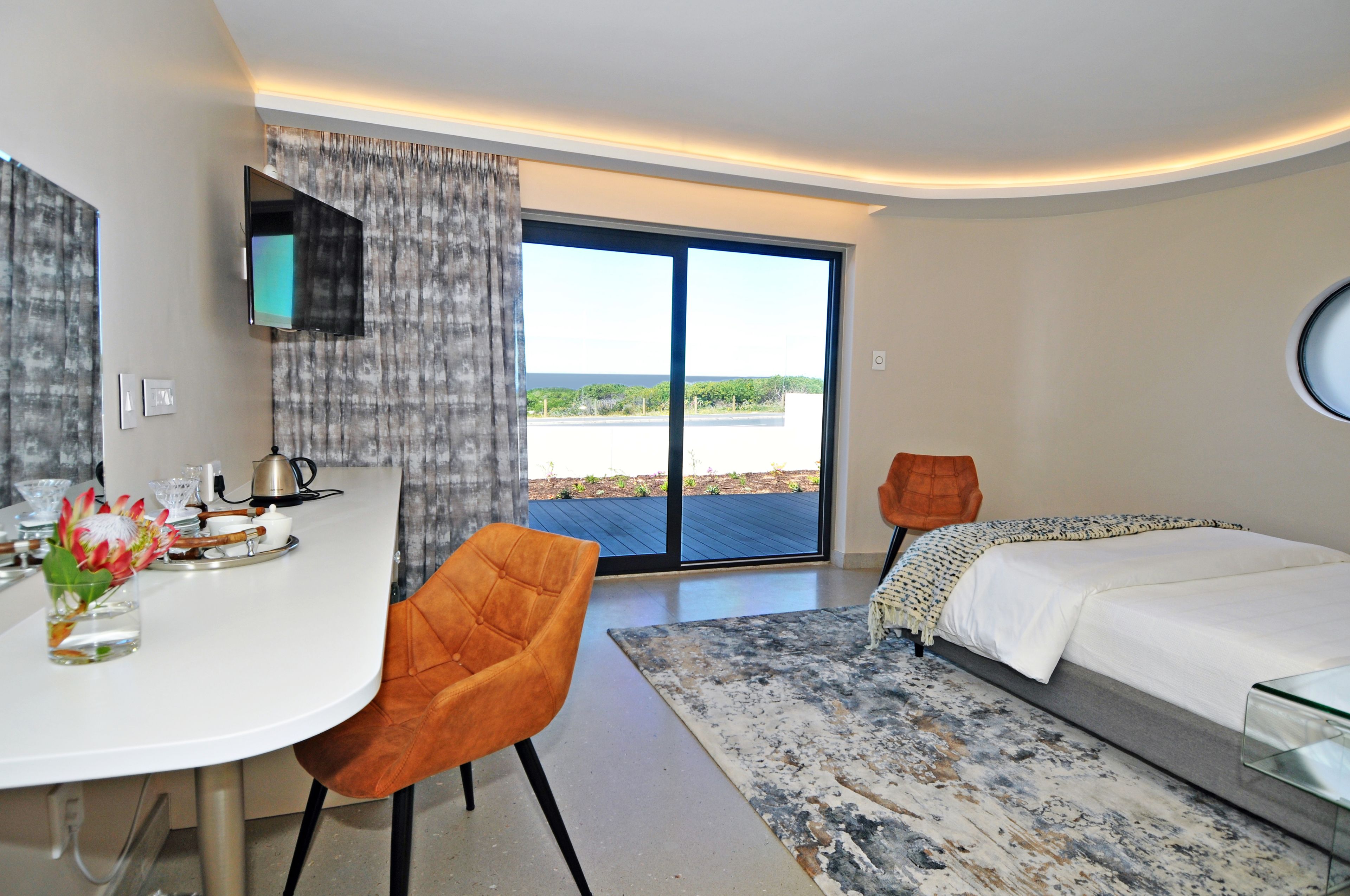 A modern hotel room with a large sliding glass door to the outside, a bed, a desk with a chair, and a small table with a vase of flowers.