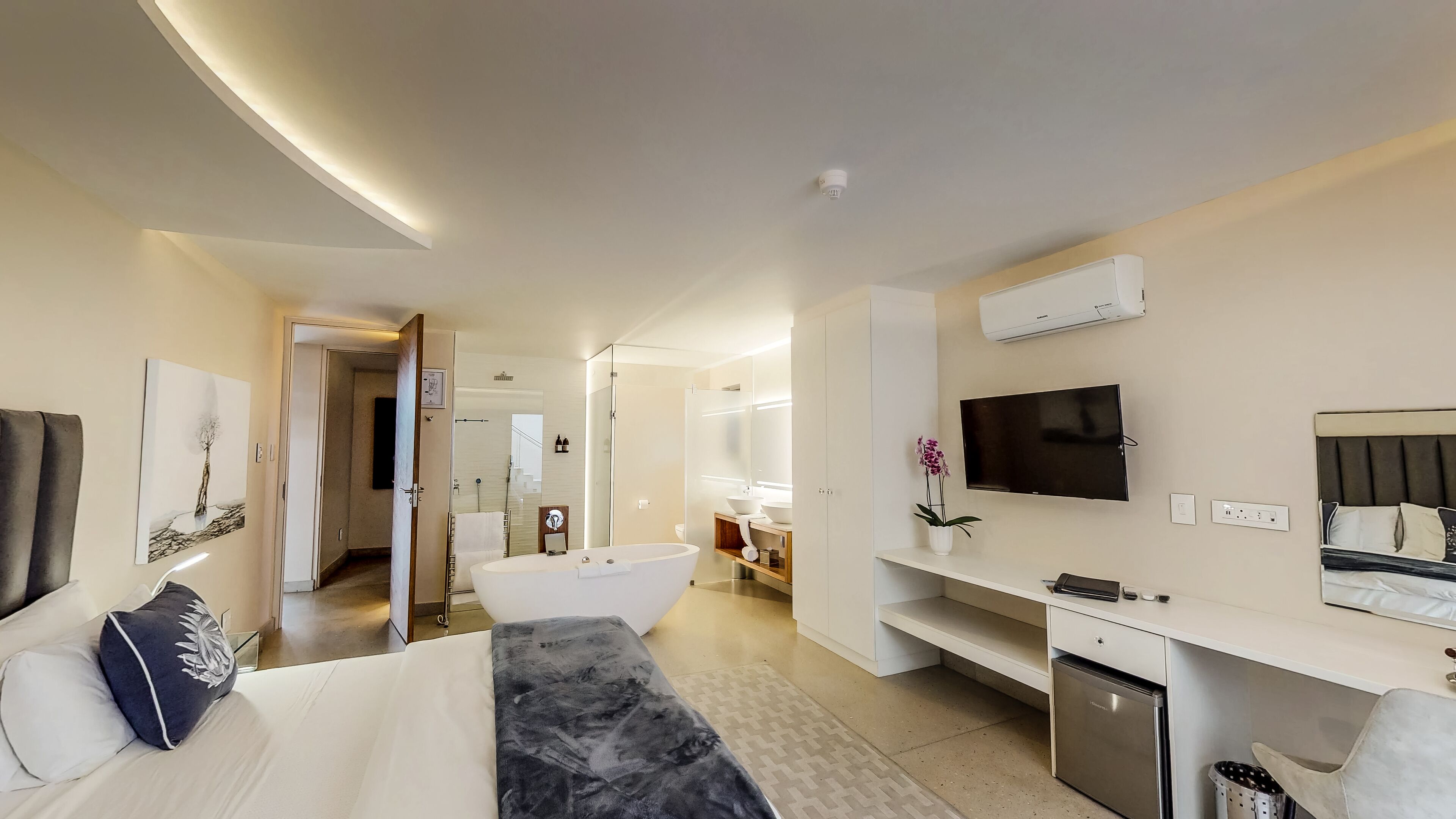 Ein modernes Hotelzimmer mit einem großen Bett, einem Fernseher, einem Schreibtisch, einer kleinen Minibar und einem Badezimmer mit Badewanne und Dusche.