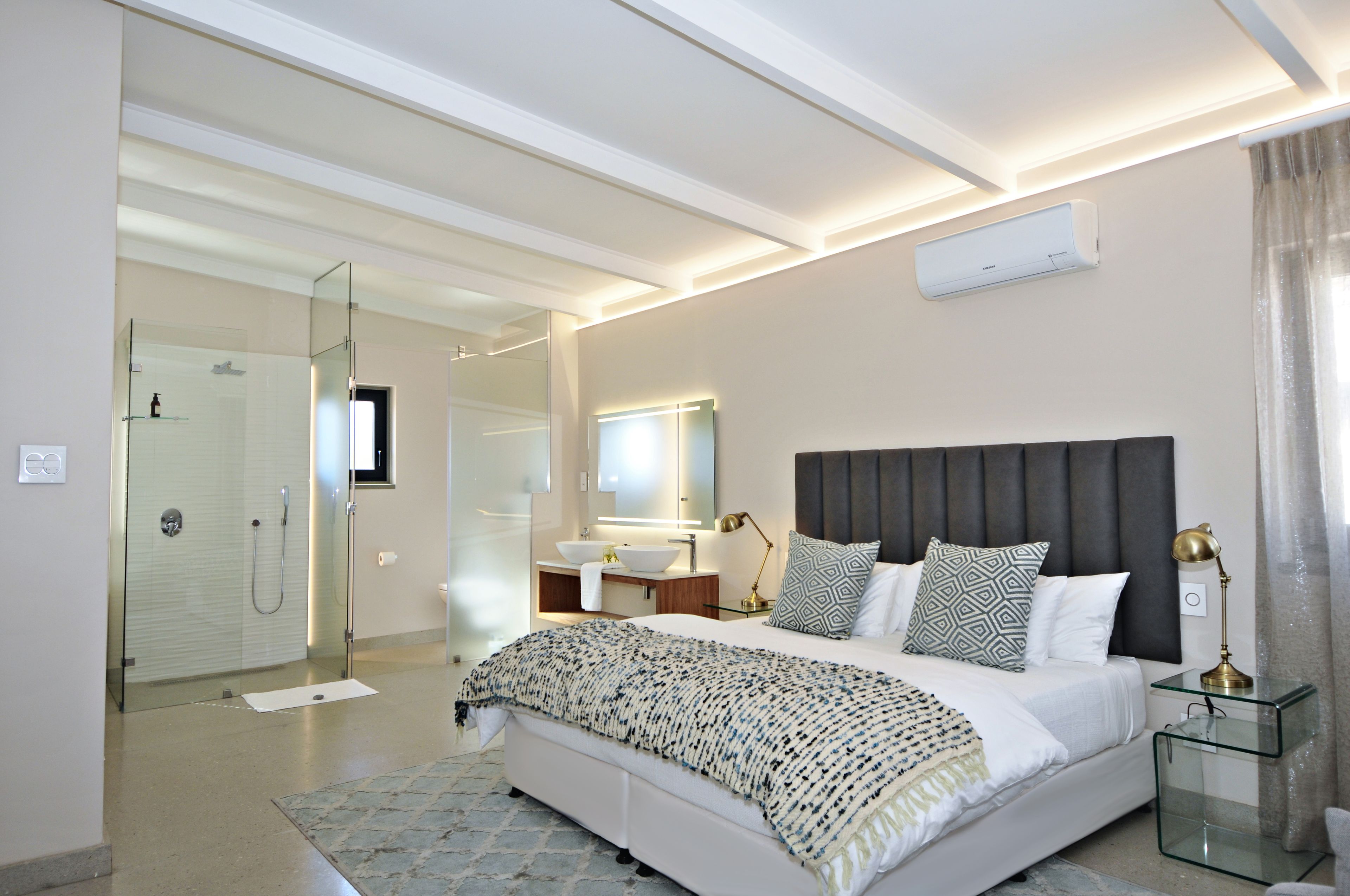 A modern bedroom with a large bed, bedside tables, a glass shower enclosure, and a bathroom with a sink and mirror.