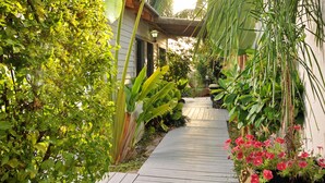 Un sendero de madera bordeado de exuberante vegetación y plantas en macetas que conduce a un edificio con tejado inclinado.