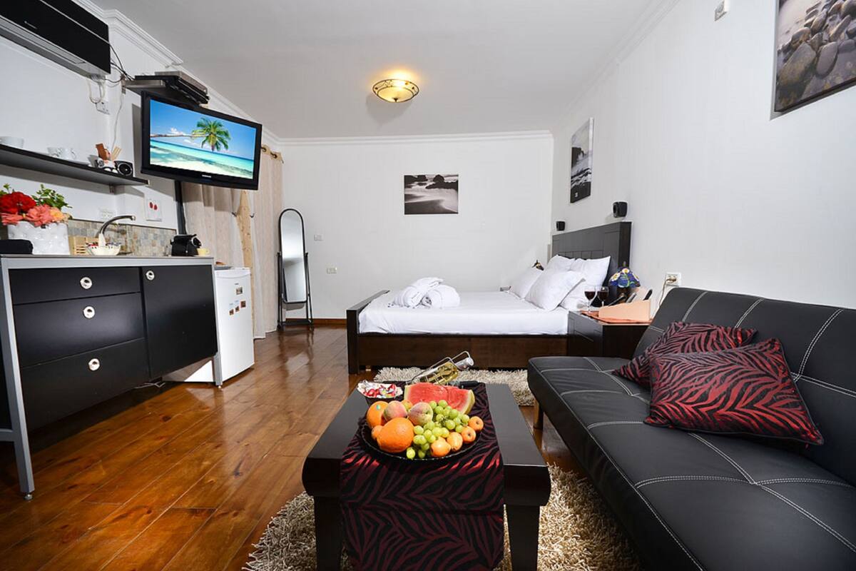A modern hotel room with a bed, a black leather sofa, a dining table with fruit, a TV, and a small kitchenette.