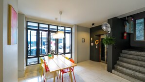 A modern dining area with a large window, a wooden table, colorful chairs, a hanging light fixture, and a staircase leading to an upper level. - Get GuestHouse 2 (Hat Yai)
