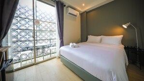 A hotel room with a bed, a desk, a chair, a balcony with a patterned railing, and an air conditioning unit. - Get GuestHouse 2 (Hat Yai)