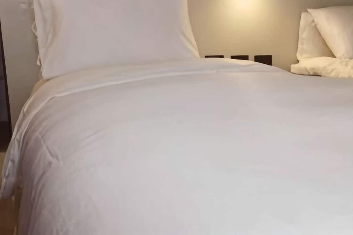Una cama con ropa de cama blanca, un cabecero y una lámpara fijada a la pared.