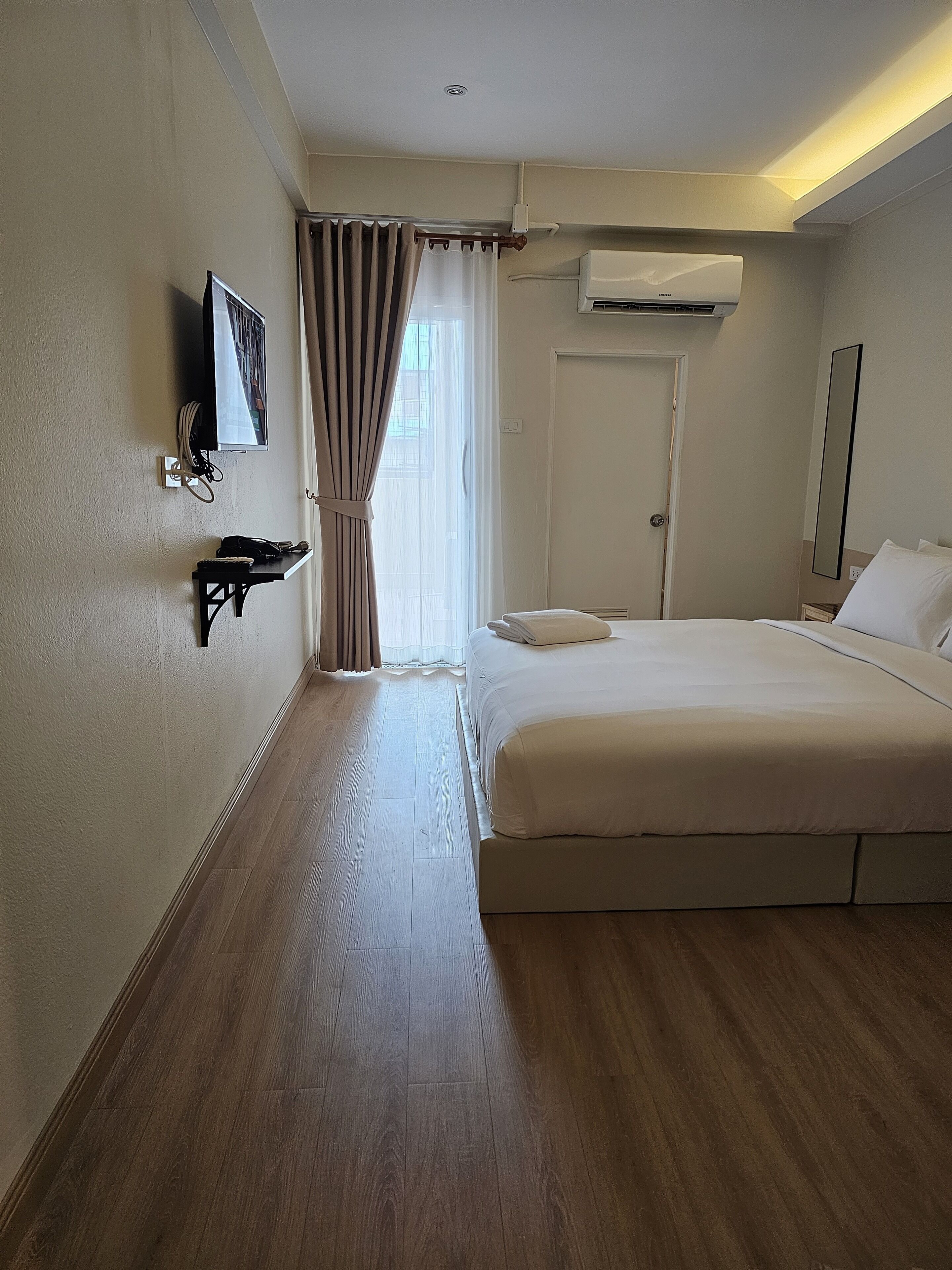 A hotel room with a bed, a flat-screen TV mounted on the wall, an air conditioning unit, a door, and a window with curtains.