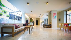 A spacious, well-lit interior with a modern design, featuring a seating area with a sofa and tables, a dining area with chairs, and a large window providing natural light. - Get GuestHouse 2 (Hat Yai)