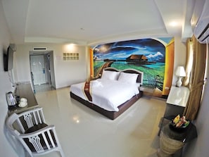A hotel room with a large bed, a desk with a chair, a television, and a mural of a coastal scene. - Bcp Hotel Rayong (Ban Chang)