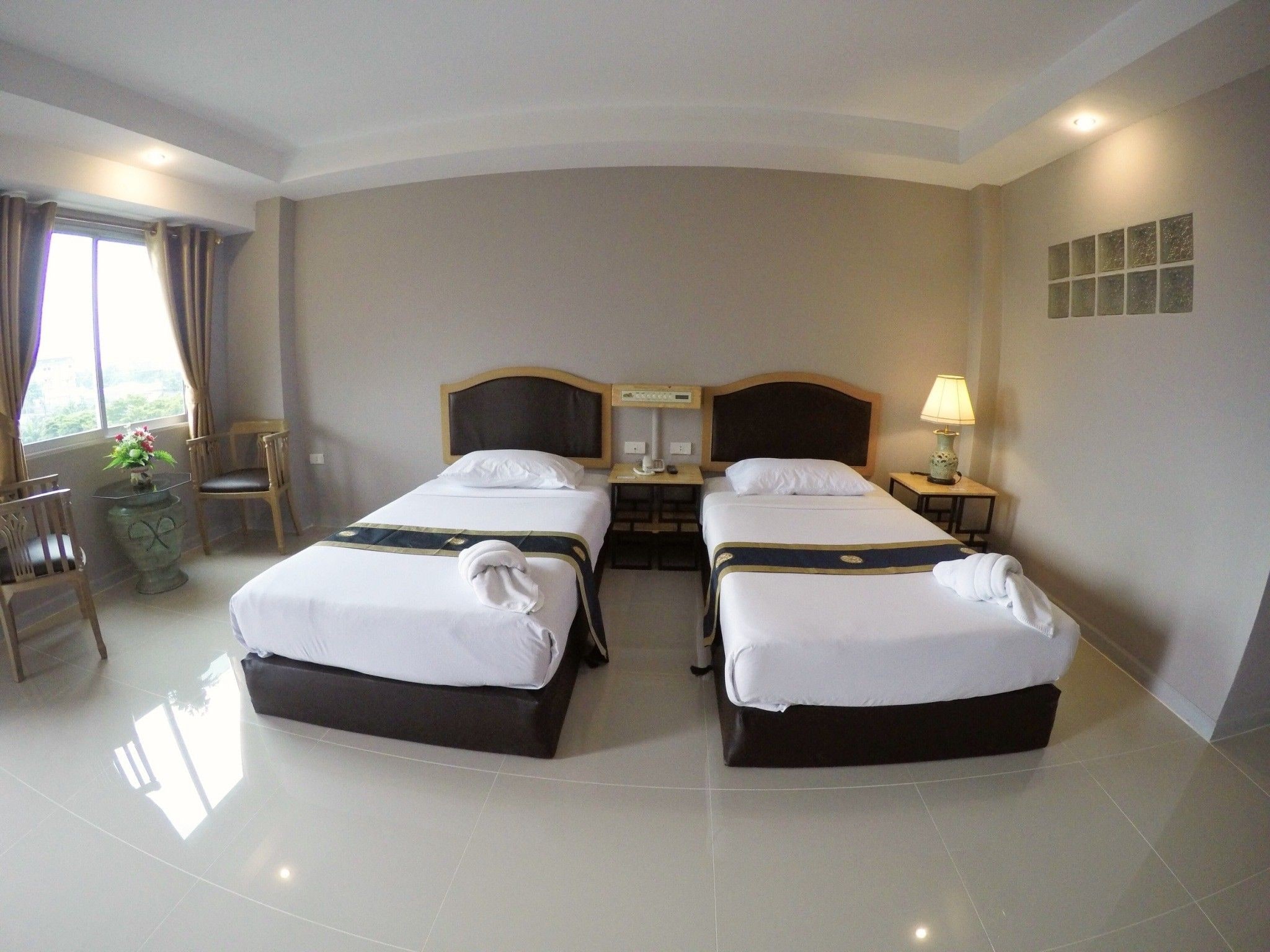 A hotel room with two beds, a small table with a lamp, a chair, and a window with curtains.