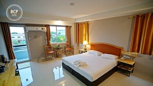 A hotel room with a bed, two chairs, a small table, a nightstand, and a window with curtains. - Bcp Hotel Rayong (Ban Chang)