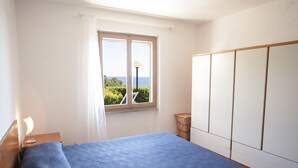 A bedroom with a bed, a window with a view, a nightstand, and a wardrobe. - Appartamento Il Maretto (Capoliveri)