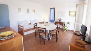 A dining area with a round table, six chairs, a sofa, a TV, and a small desk with a lamp. - Appartamento Il Maretto (Capoliveri)