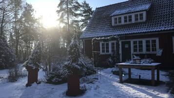 Ein mit Schnee bedecktes Haus mit Ziegeldach, umgeben von schneebedeckten Bäumen und wolkenlosem Himmel.