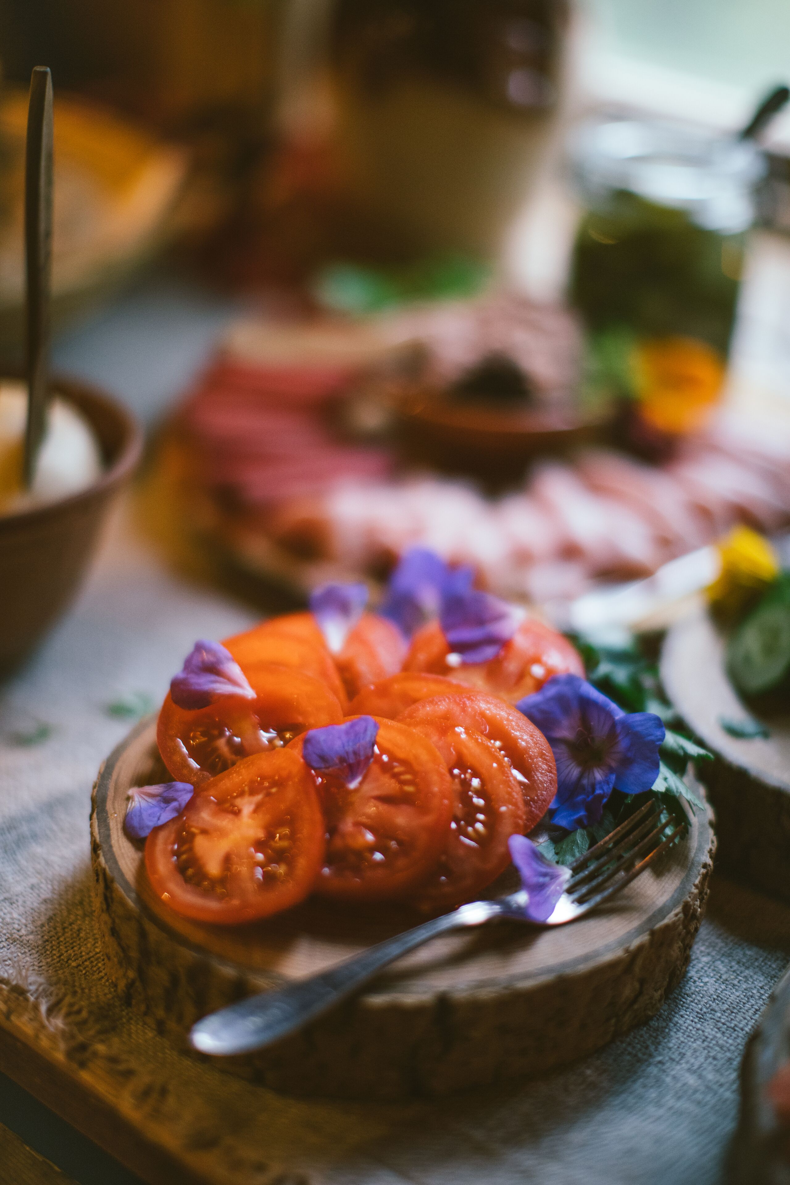 Ein Holzboden mit Tomatenscheiben und violetten Blüten, eine Gabel und ein unscharfer Hintergrund mit weiteren Speisen.
