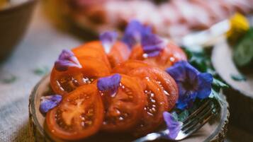 Ein Holzboden mit Tomatenscheiben und violetten Blüten, eine Gabel und ein unscharfer Hintergrund mit weiteren Speisen.
