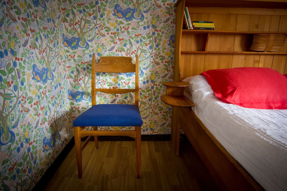 A room with a wooden chair with a blue cushion, a wooden bed with a red pillow, and a bookshelf with books and a plate rack.