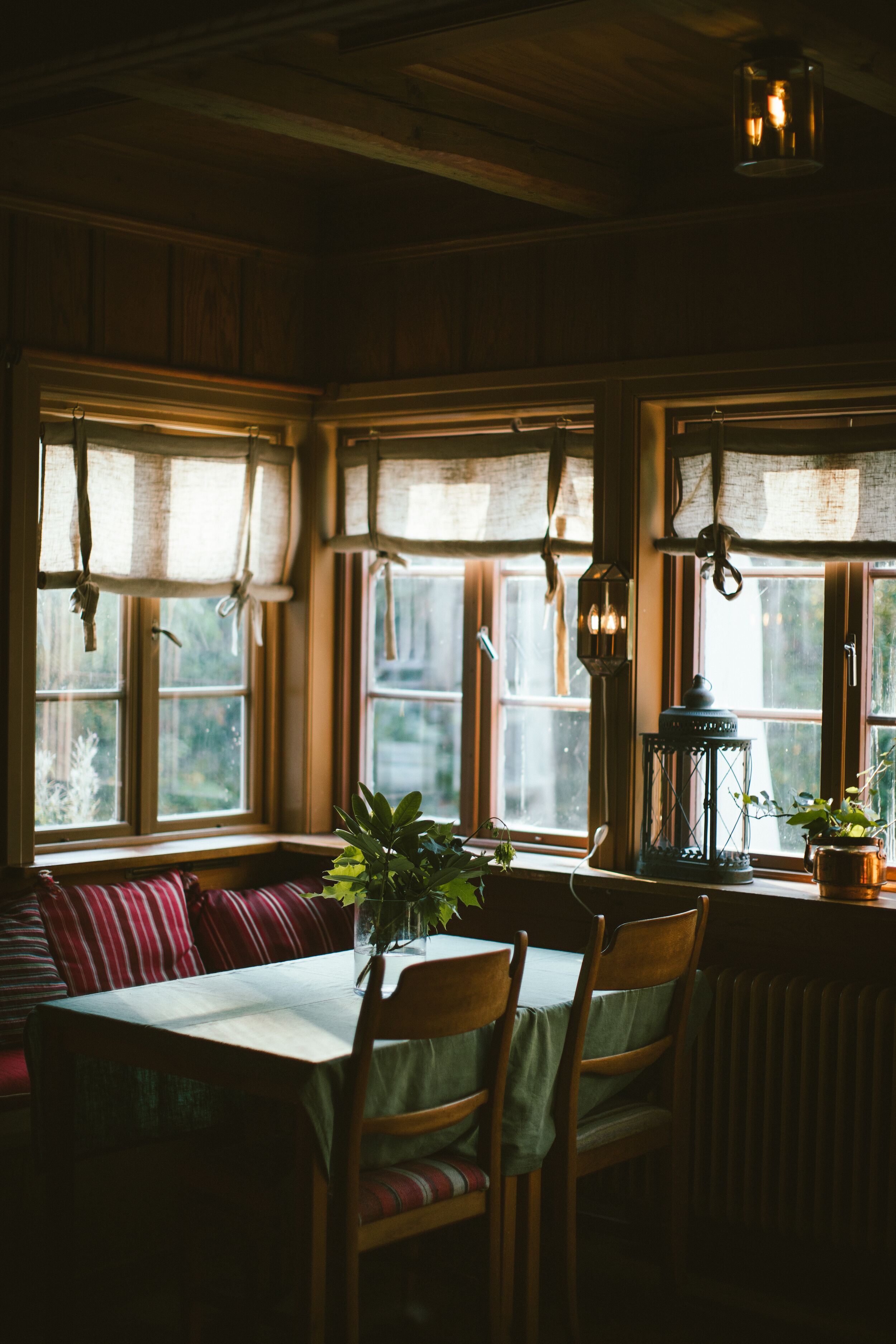 A cozy dining area with a wooden table, chairs, and a bench. There are windows with curtains and a lantern hanging above.