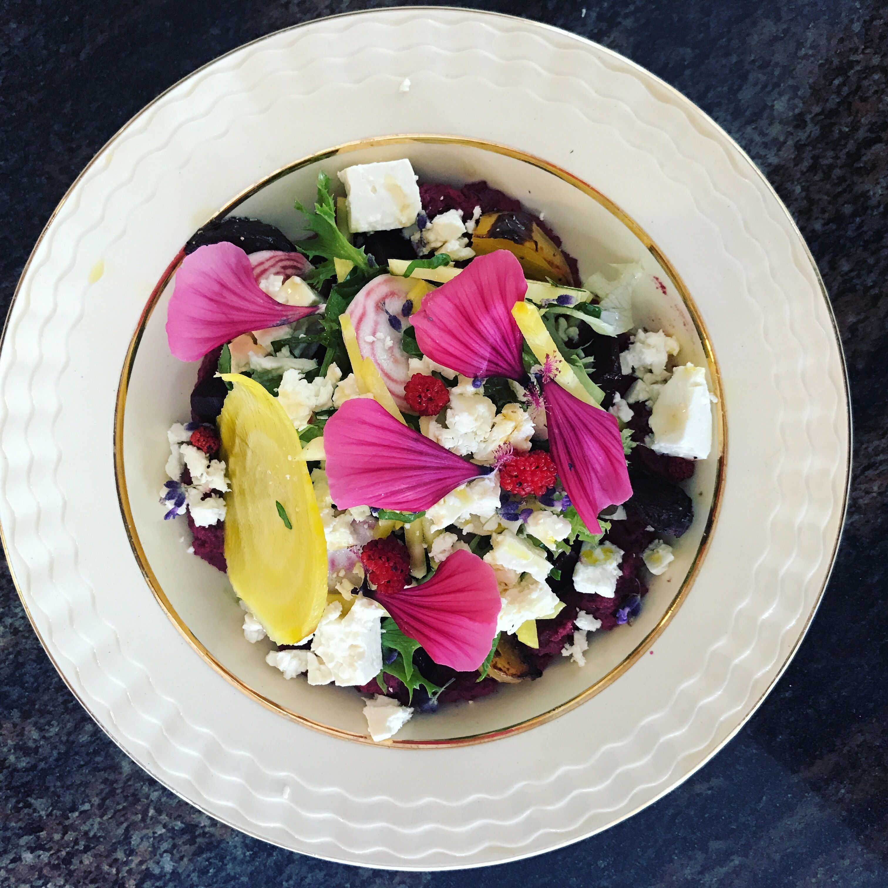 Un tazón blanco con borde dorado que contiene una ensalada con queso feta, bayas rojas y flores rosas.