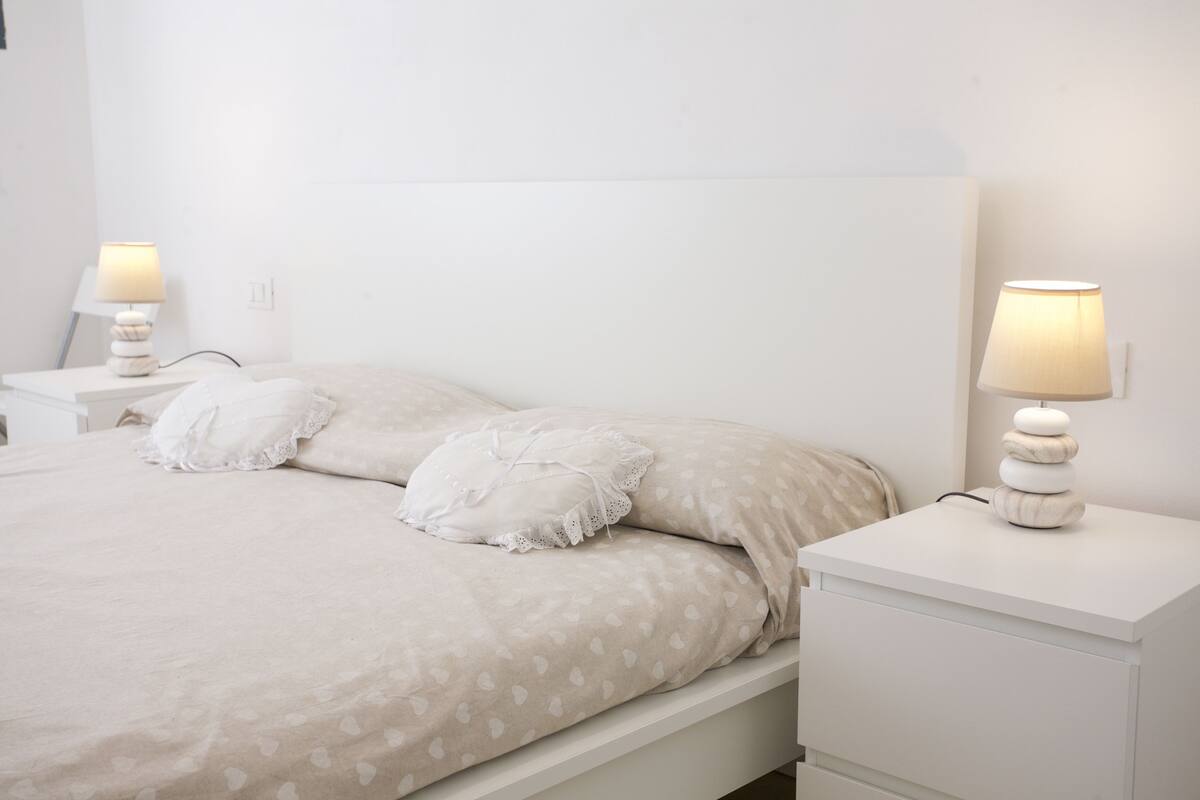 A neatly made bed with a heart-patterned bedspread, two bedside lamps, and a white nightstand with a drawer.