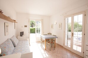 A room with a sofa, a dining table with chairs, a television on a small table, and a door leading outside. - Appartamenti Villa Anna (Capoliveri)