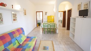 A living room with a colorful sofa, a dining area with a table and chairs, and a TV on a cabinet. - Appartamenti Arco Bianco (Capoliveri)