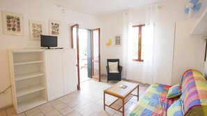 A bright living room with a colorful sofa, a wooden table, a TV on a stand, and a chair. There are framed pictures on the wall and a door leading outside. - Appartamenti Arco Bianco (Capoliveri)