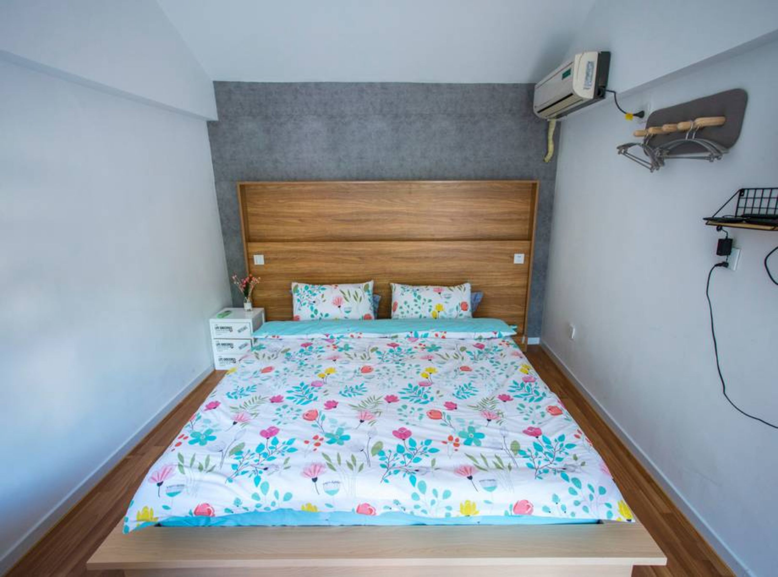 A bedroom with a wooden headboard, floral bedding, a nightstand, and an air conditioning unit on the wall.