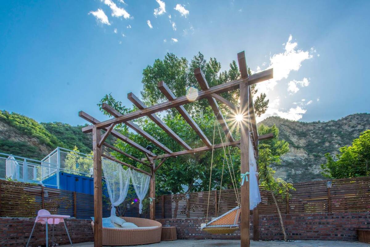 A wooden pergola structure with a swing and a cushioned seating area, set against a backdrop of a mountainous landscape.