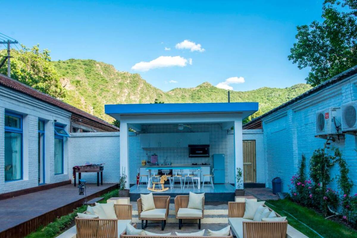 A modern outdoor kitchen area with a bar and seating, surrounded by a white building and lush greenery.