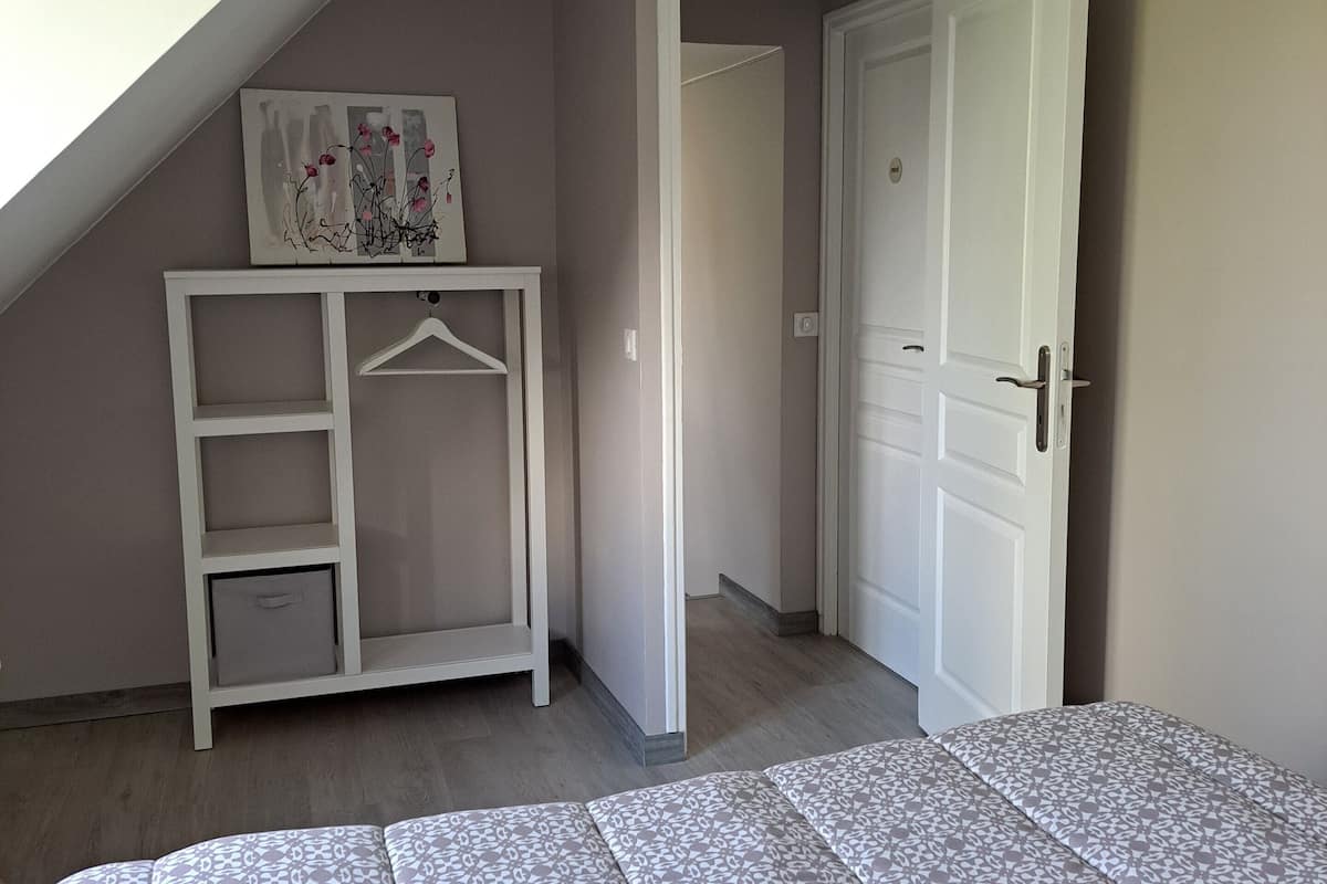 Ein Schlafzimmer mit schrägem Dach, einem Bett mit gemusterten Betttextilien, einem weißen Kleiderschrank und einer Tür zu einem anderen Raum.