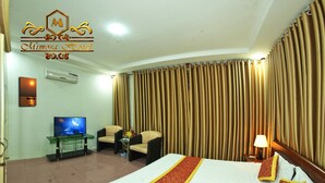 Habitación de hotel con una cama grande, un televisor sobre una mesa de cristal, dos sillones, una mesita con un jarrón de flores y un telón de fondo.