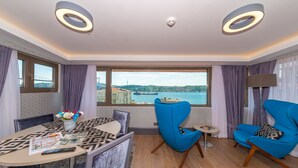 Una camera d'albergo moderna con angolo ristoro, due poltrone blu, un tavolino con un vaso di fiori e un'ampia finestra che si affaccia su un ponte e sull'acqua.