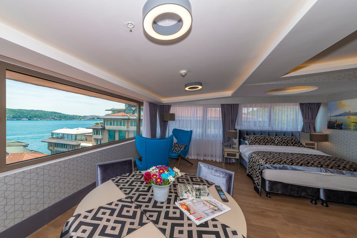 Une chambre d’hôtel moderne dotée d’une grande fenêtre donnant sur la mer et les bâtiments, d’un coin repas avec une table à motifs et d’un lit avec une couette à motifs géométriques.